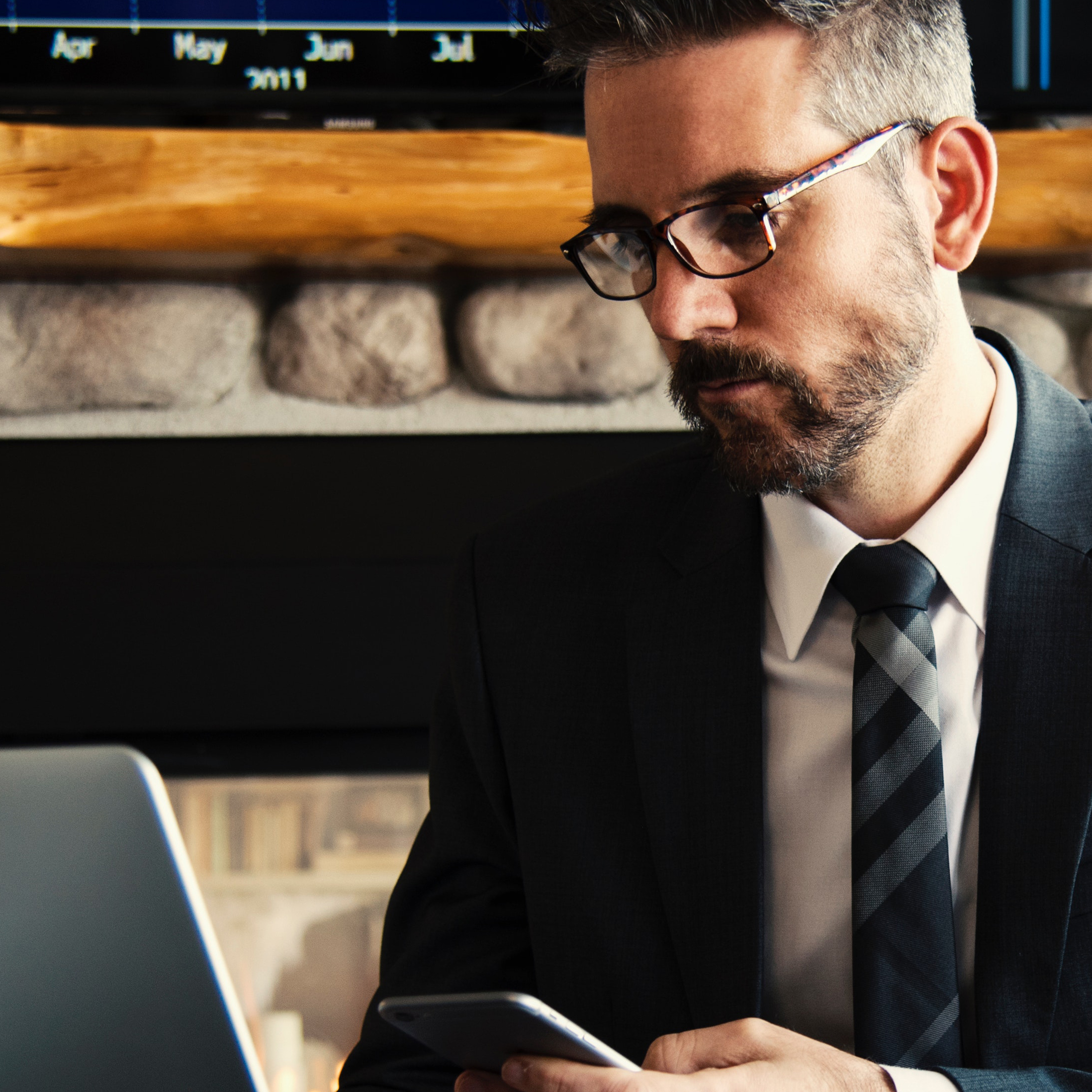 A man with glasses wearing a suit holding a phone and lookng at laptop